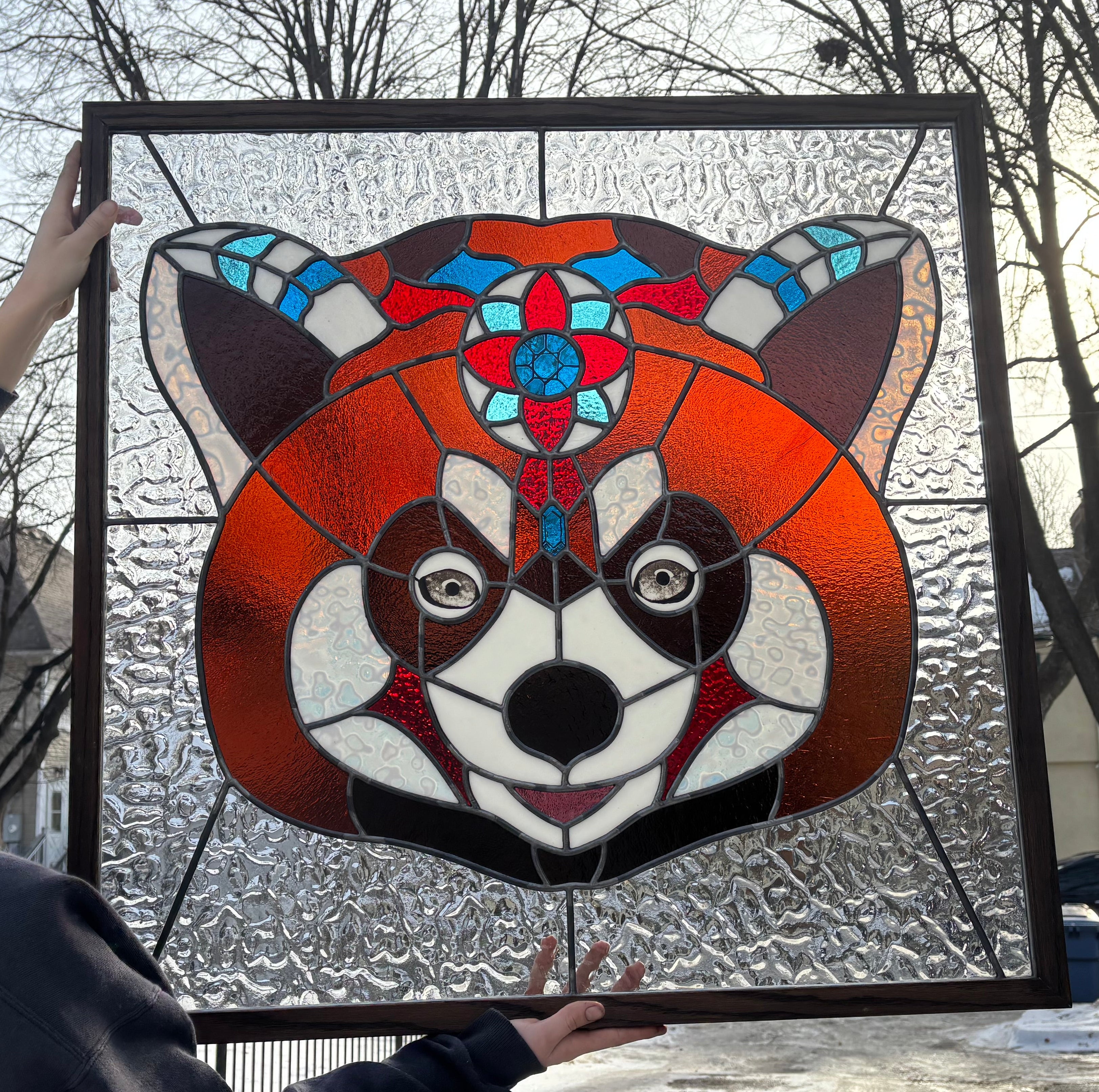 Person holding a stained glass panel of a red panda's face outdoors on a snowy day.