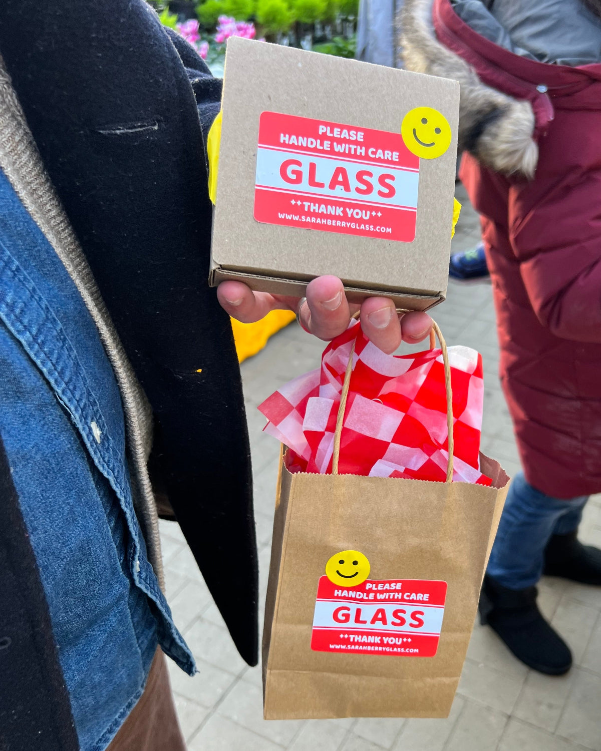 A person holding two brown paper gift boxes with red and white 'GLASS' handling stickers and a red-and-white checkered tissue pouch, photographed outdoors with other people in winter clothing in the background.